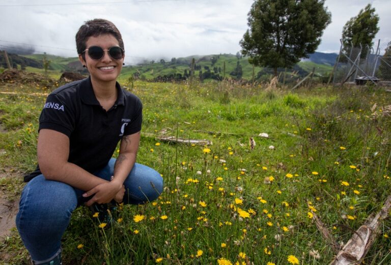 Vanesa smiling to the camera on a field trip, wearing her Press shirt. In the background, mountains and yellow flowers.