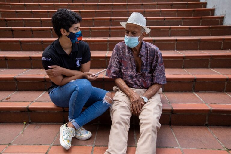 Vanesa and an old man sitting on the stairs of a church. Vanesa is holding an audio recorder close to the man.