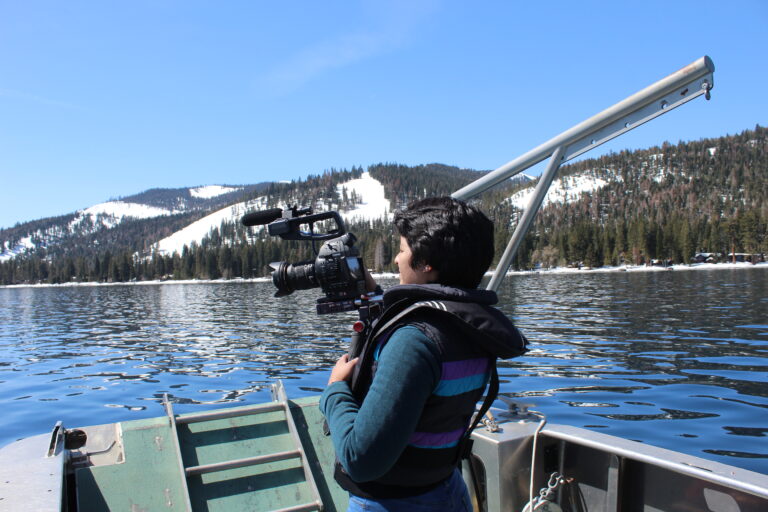 Vanesa is in a boat in Lake Tahoe, with a camera in her hands, recording the lake and the snowy mountains in the background.